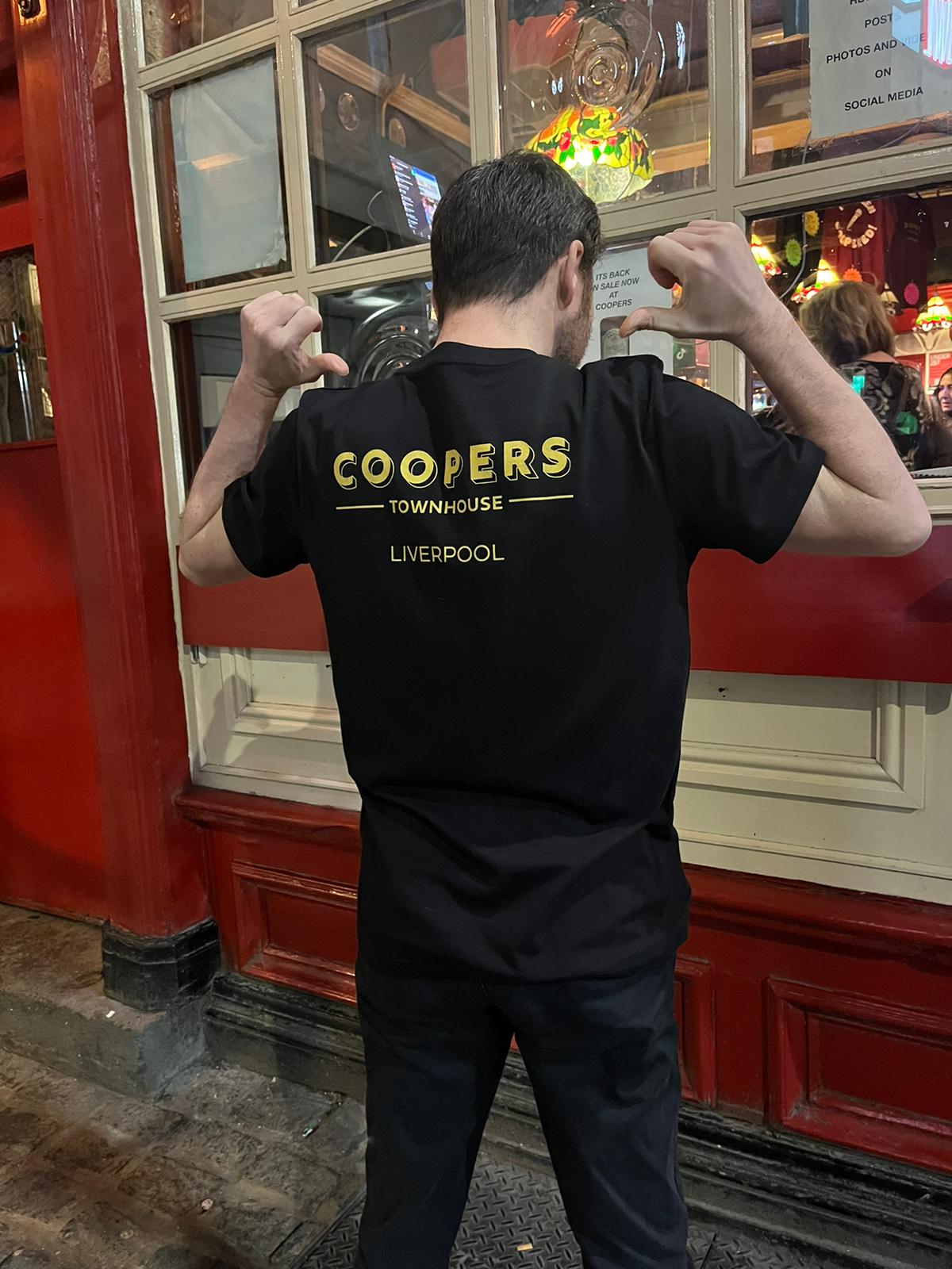 A man standing outside Coopers Pub, facing away from the camera, pointing at the back of his shirt to highlight the logo of the merchandise.